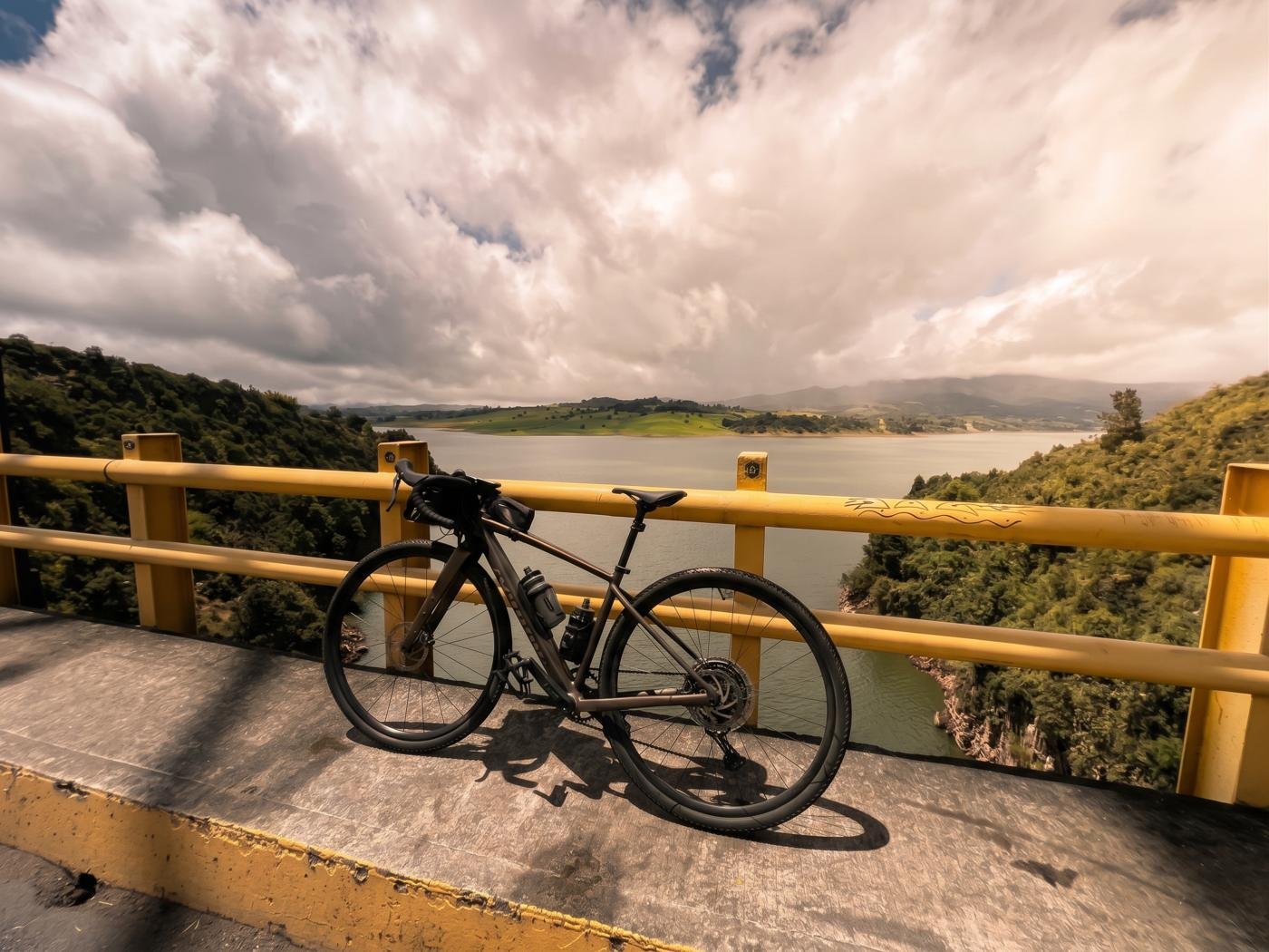 Bicicleta gravel apoyada en la baranda amarilla de la presa del Sisga, con el embalse y las montañas al fondo